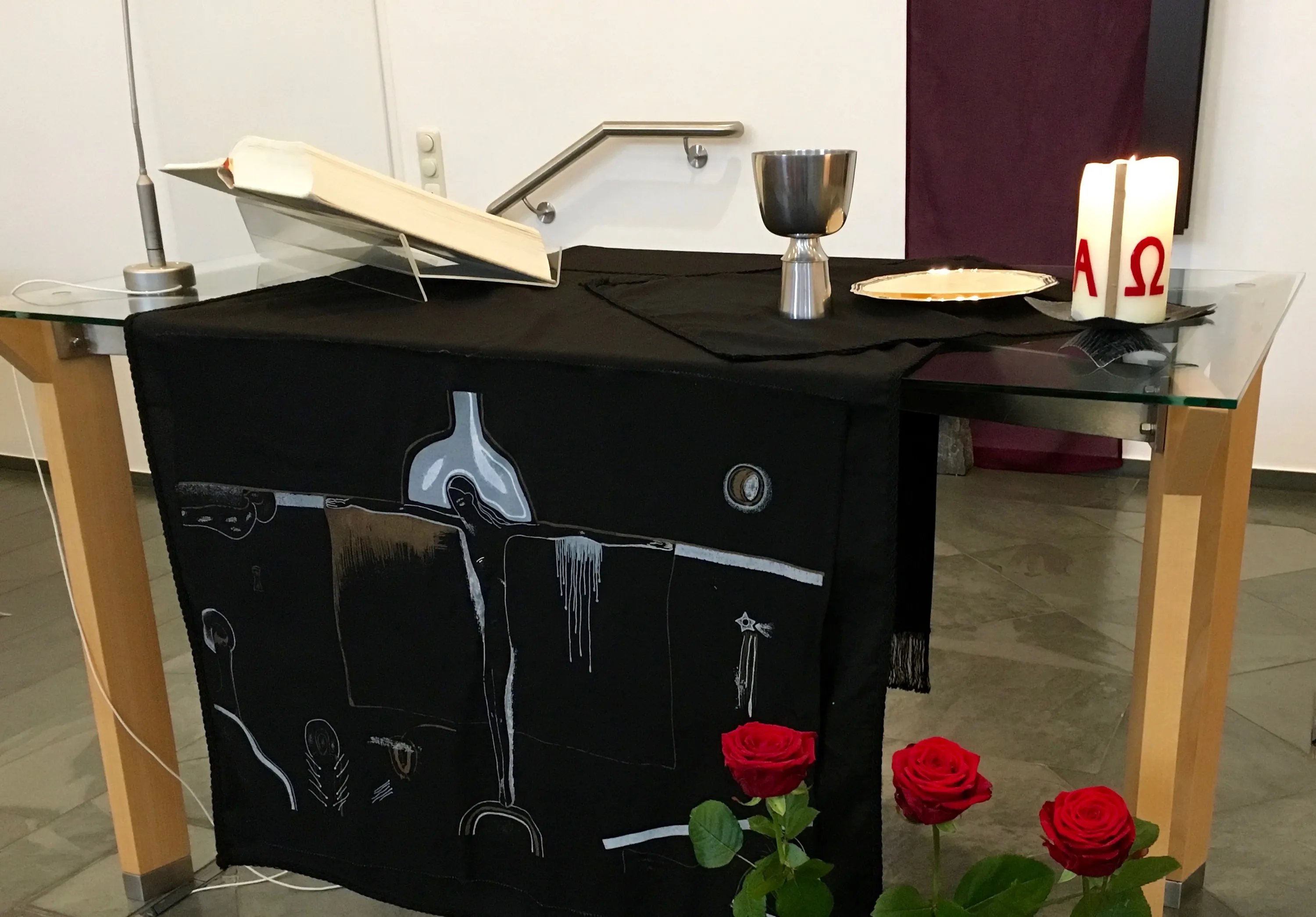 Abendmahlstisch mit Abendmahl, Kerze, Bibel – schwarze Paramente mit dem Gekreuzigten – davor drei rote Rosen