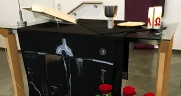 Abendmahlstisch mit Abendmahl, Kerze, Bibel – schwarze Paramente mit dem Gekreuzigten – davor drei rote Rosen